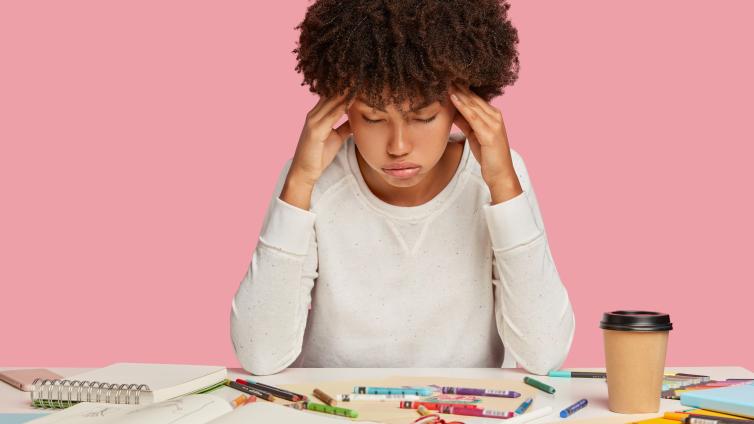 Stressed student with hands on her temples against pink background and sitting at a messy table with books, crayons, and a coffee cup.